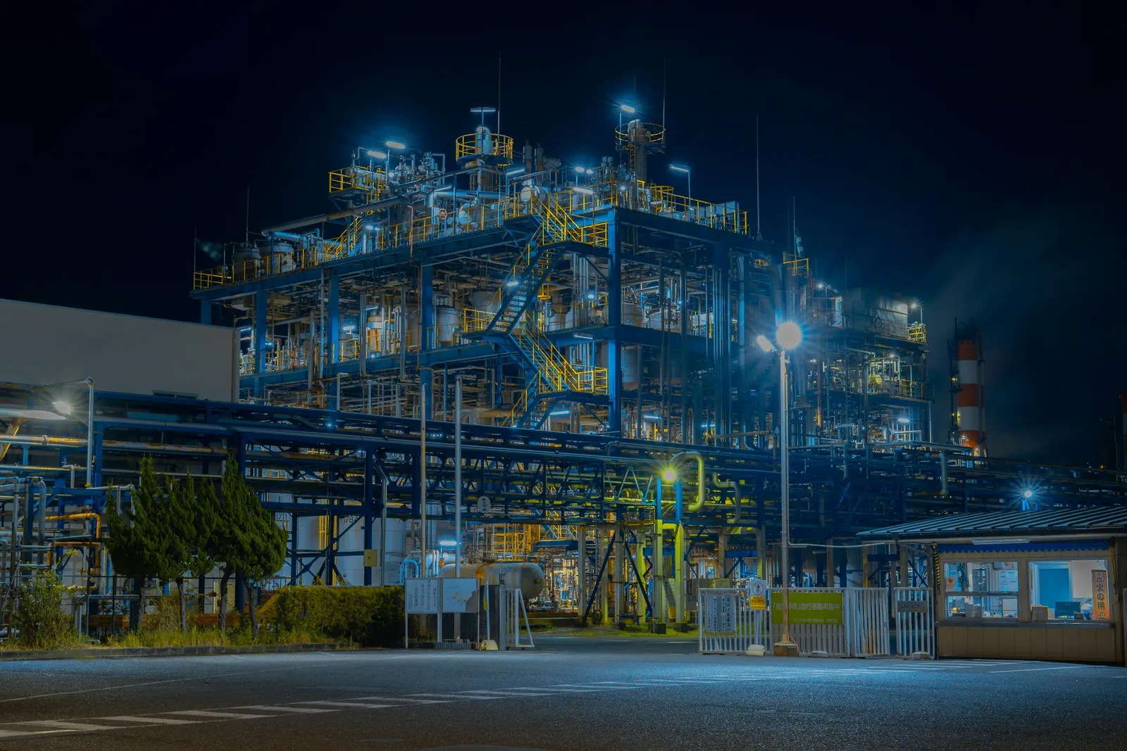 Industrial facility lit up at night, seen from behind its perimeter fence and gate